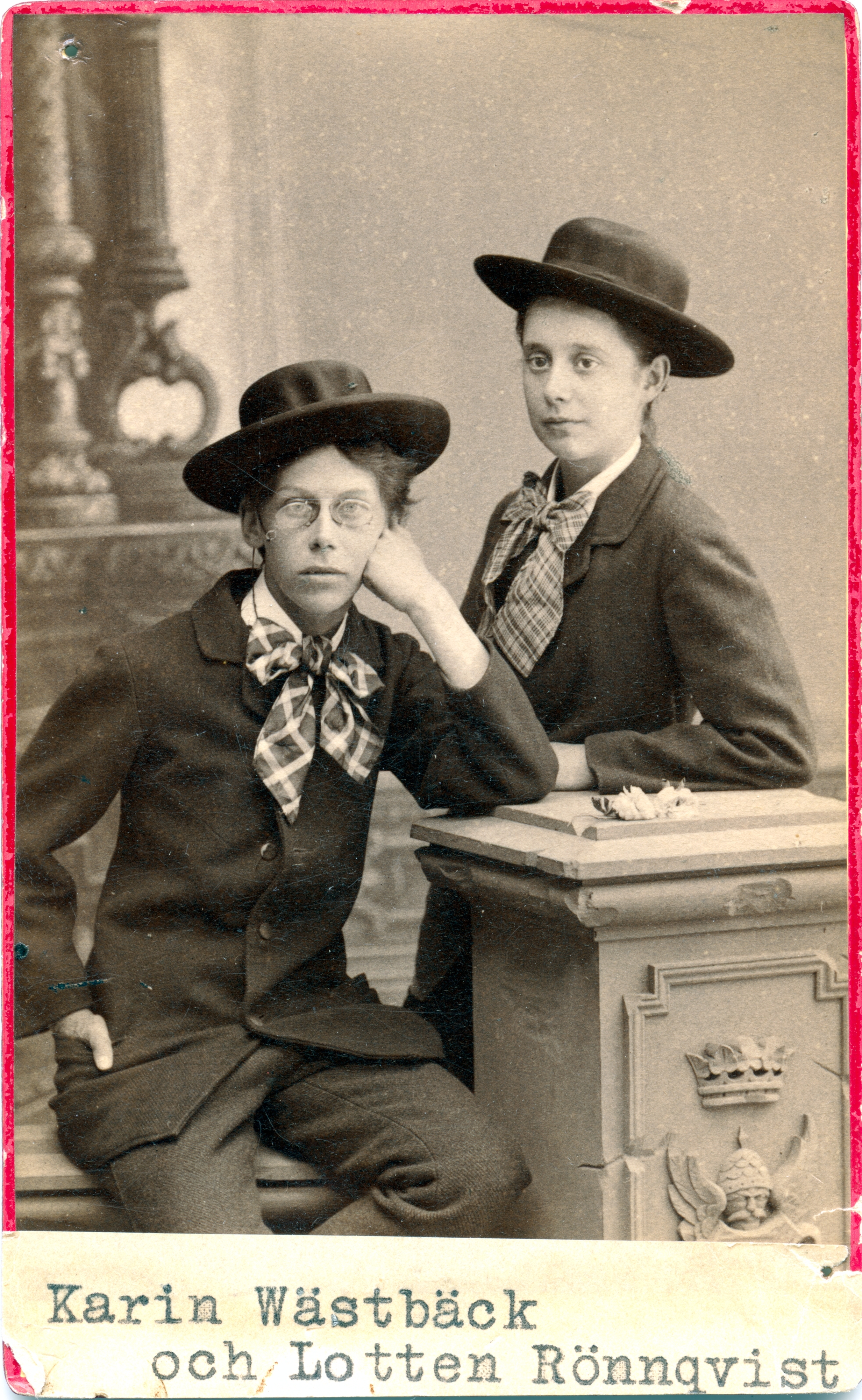 svartvitt foto. Två kvinnor i 20 års åldern. De är klädda i herrkostym och herr hatt med stora mönstrade flugor. En av kvinnorna sitter med kroppen och blicken vänd mot kameran. Den andra står med kroppen och vikten riktad mot vänster.